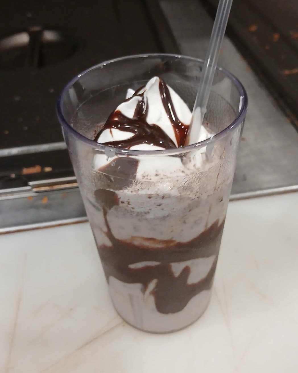 A thick milkshake in a tall cup, topped with whipped cream and chocolate syrup, with a straw