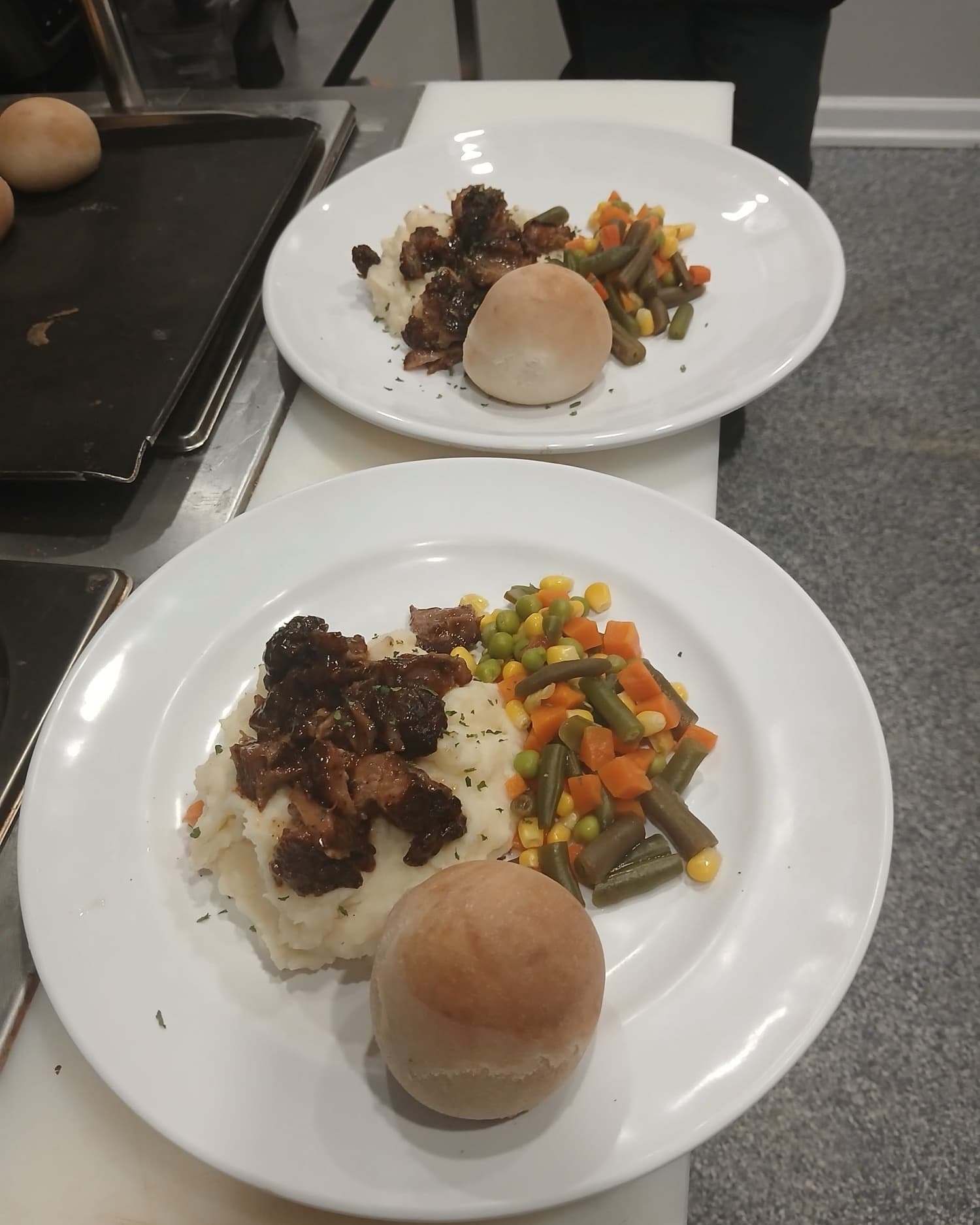 Two plates of roast over mashed potatoes with mixed vegetables and a dinner roll, ready to serve